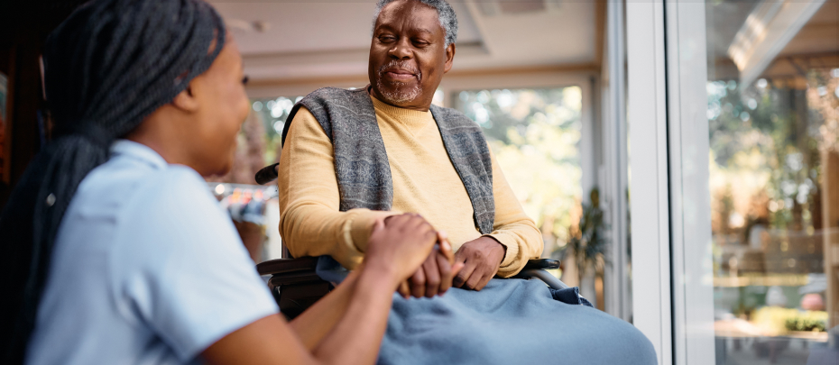 An elderly woman in a wheelchair holds hands with a young woman, symbolizing connection and support between generations.
