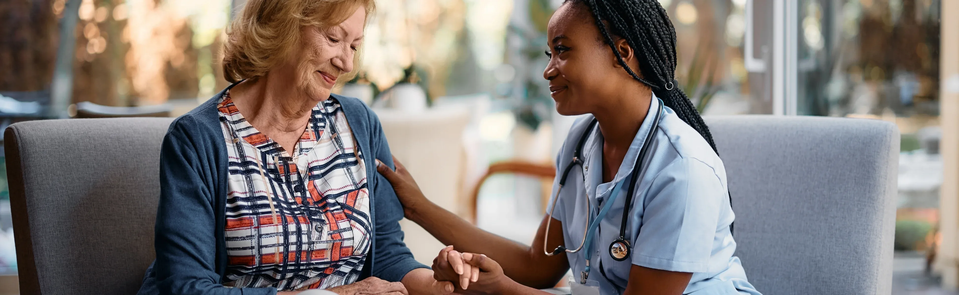 A nurse and a patient are seated together on a couch, engaged in a supportive conversation in a comfortable setting.