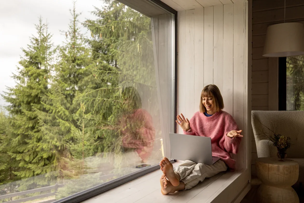 A woman sits on a window sill, focused on her laptop, with natural light streaming in around her.