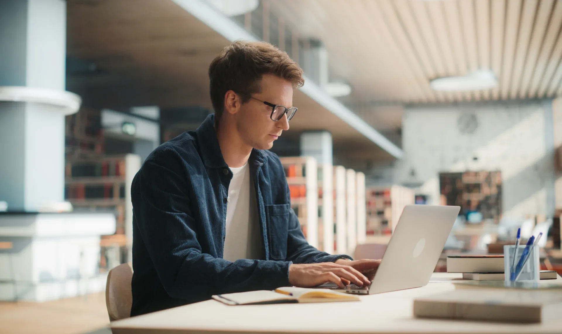 A man wearing glasses sits at a table, focused on his laptop, engaged in work or study.