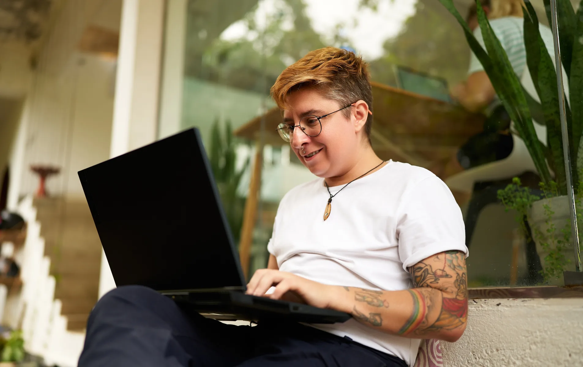 A tattooed man sits on a bench, focused on his laptop, surrounded by a serene outdoor environment.