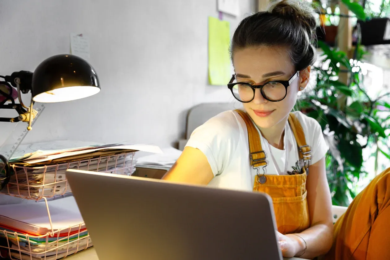 A woman wearing glasses is focused on her laptop, engaged in work at a well-lit desk.