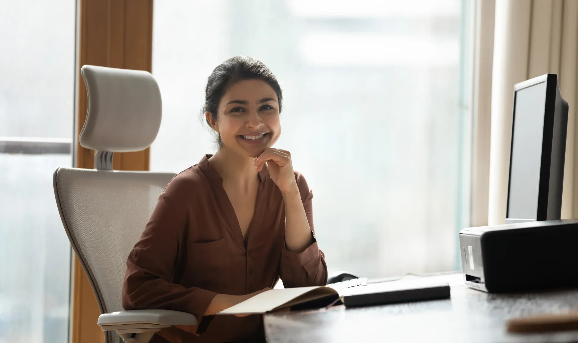 A woman focused on her laptop while seated at a desk, surrounded by a tidy workspace and natural light.