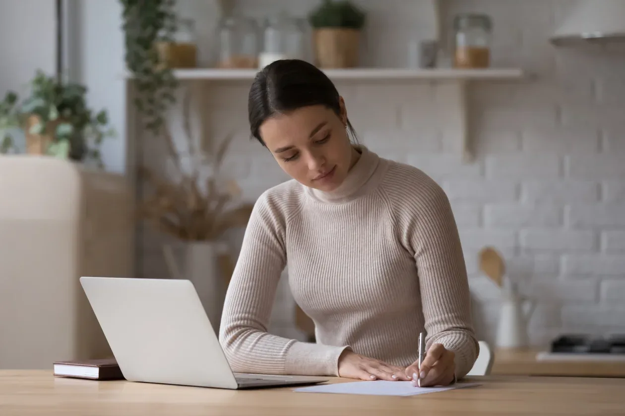 A woman sits at a table with a laptop, writing on a piece of paper, focused and engaged in her task.